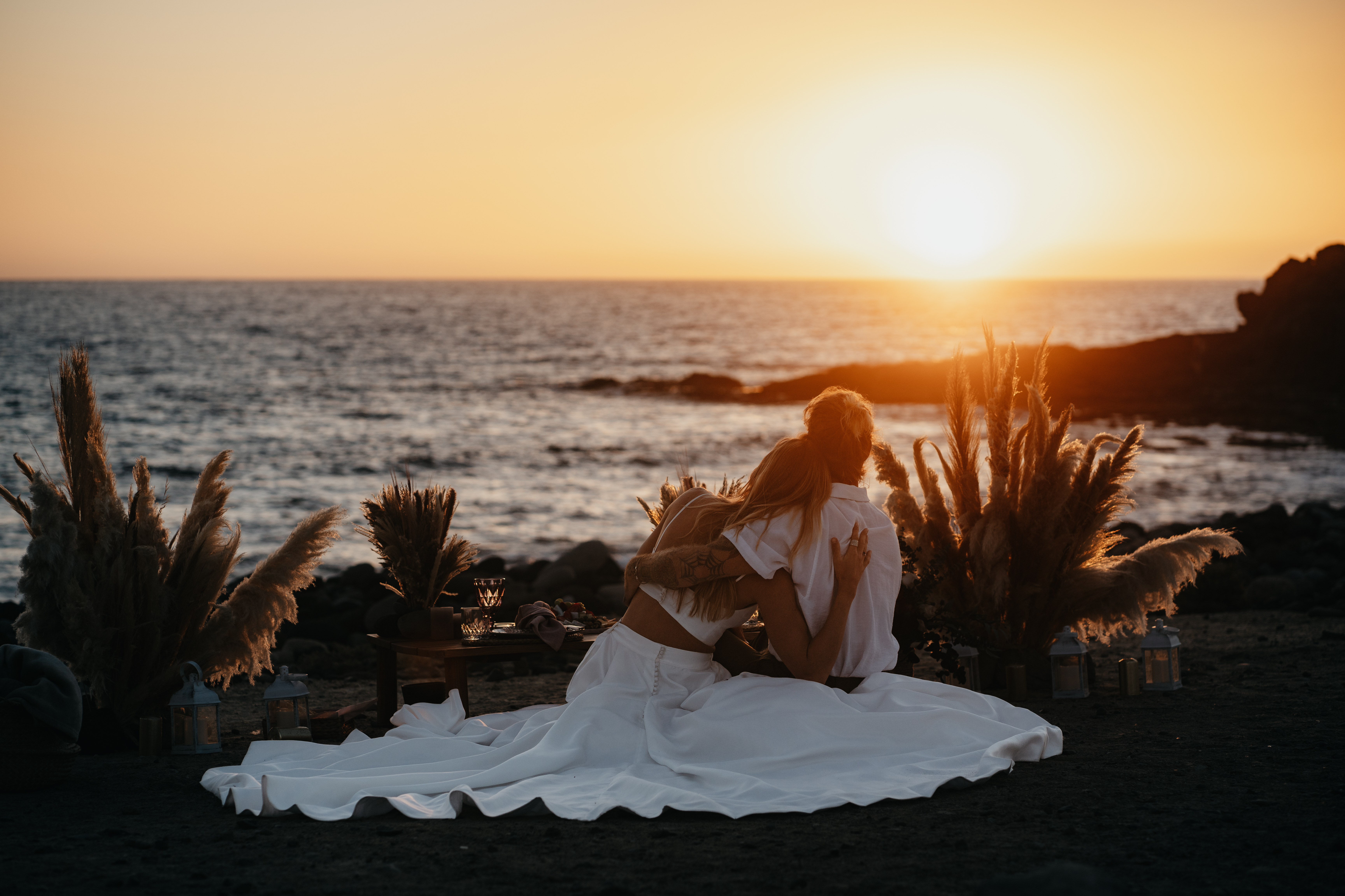 Un couple de mariés en tenue estivale assis bras dessus bras dessous sur la plage, observant le coucher du soleil.